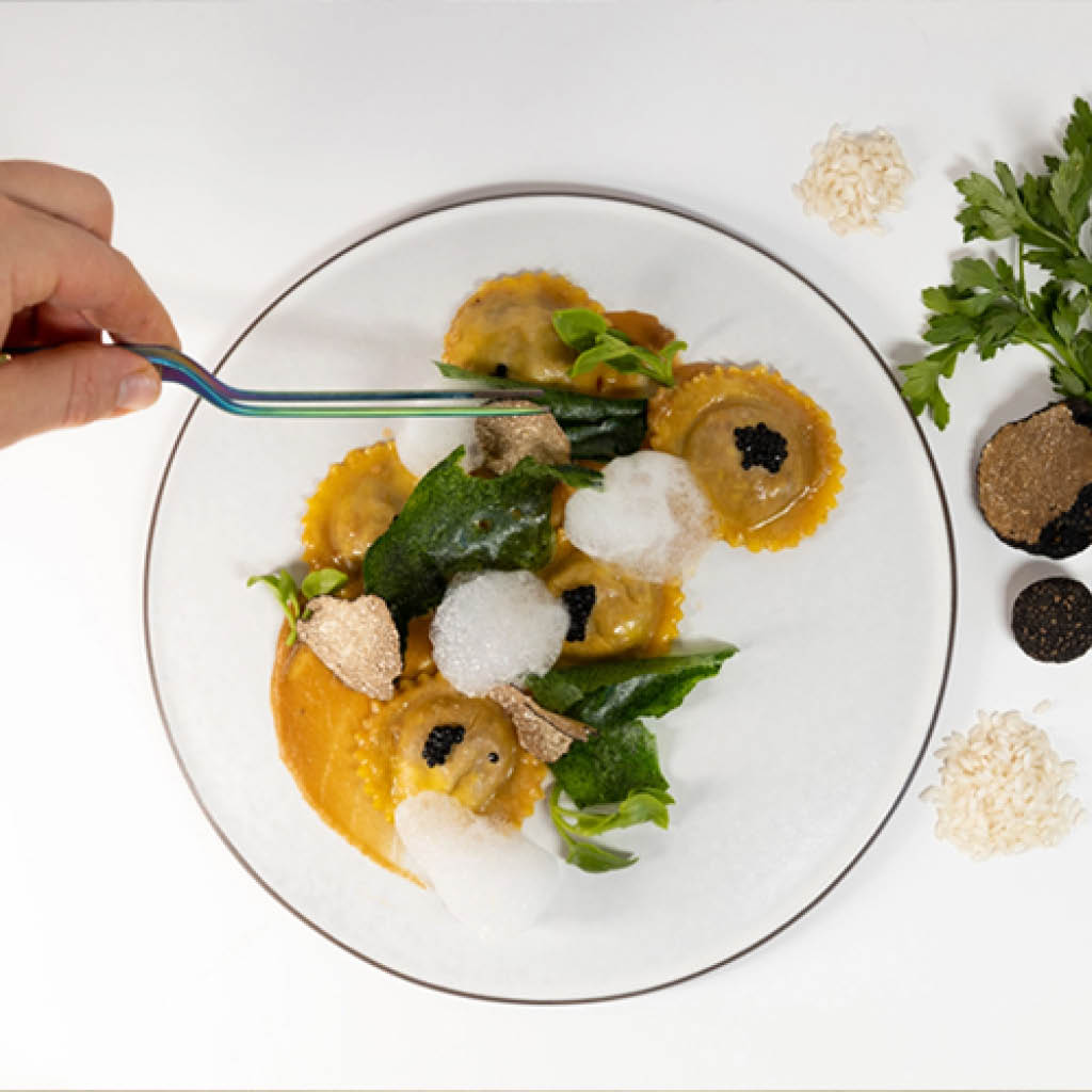 Ein Teller mit kunstvoll angerichteten Ravioli, verfeinert mit Trüffeln, Blattspinat und Schaum. Eine Hand richtet das Gericht mit einer Pinzette präzise an. Dieses Gourmetgericht repräsentiert das gehobene kulinarische Erlebnis, das in einem exklusiven Golfhotel oder Fine-Dining-Restaurant angeboten wird.