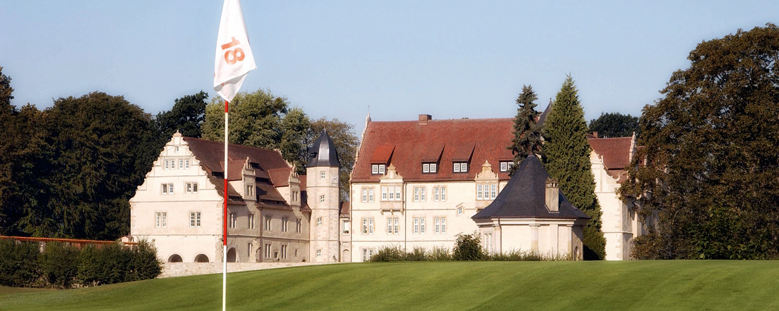 Ein Golfplatz mit einer Fahne an Loch 18 erstreckt sich vor einem historischen Schlossgebäude. Diese malerische Kulisse kombiniert die Eleganz eines Schlosses mit der Schönheit einer erstklassigen Golfanlage und bietet Gästen eines exklusiven Golfhotels ein unvergleichliches Erlebnis.