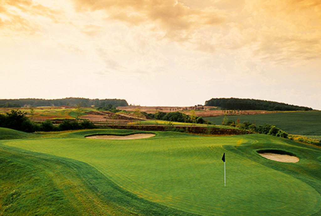 Weitläufiger Golfplatz bei Sonnenuntergang mit perfekt gepflegtem Grün, umgeben von sanften Hügeln und Bunkern. Diese malerische Landschaft betont die Schönheit und Ruhe der Golfanlage, ideal für einen Aufenthalt in einem exklusiven Golfhotel.