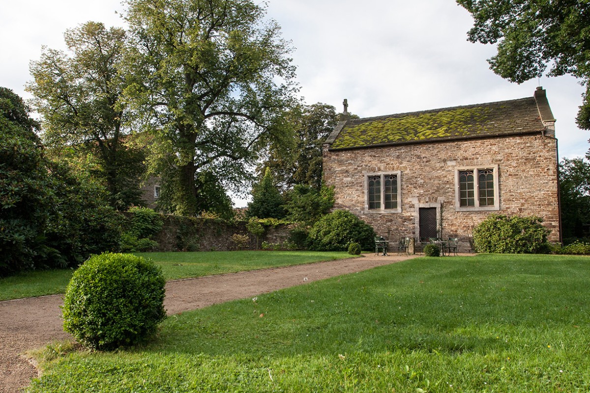 Charmantes, historisches Steingebäude inmitten einer grünen Gartenanlage, ideal für eine intime Hochzeitszeremonie oder eine romantische Hochzeitslocation im Freien. Die Umgebung ist von großen Bäumen und gepflegtem Rasen umgeben und bietet eine ruhige, malerische Kulisse für Hochzeitsfotos.