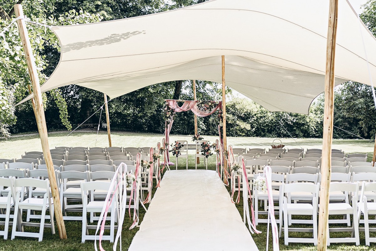 ind in Reihen aufgestellt, und ein Blumengesteck schmückt den Altarbereich. Rosa Bänder und Blumen sorgen für eine romantische und elegante Dekoration, ideal für eine Outdoor-Hochzeit.