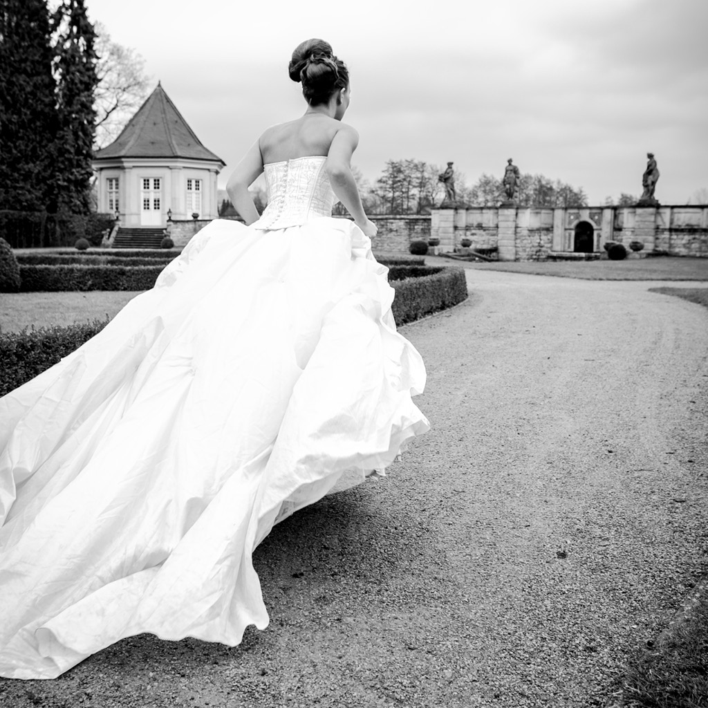 Schwarz-Weiß-Fotografie einer Braut in einem voluminösen Hochzeitskleid, die durch eine historische Gartenanlage läuft. Im Hintergrund sind ein Pavillon und kunstvolle Statuen zu sehen, was der Szene eine romantische und elegante Atmosphäre verleiht, ideal für eine Hochzeitslocation im Freien.