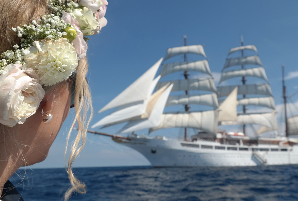 Eine Braut mit einem Blumenkranz im Haar blickt auf ein majestätisches Segelschiff, das auf dem offenen Meer segelt. Diese einzigartige Hochzeitslocation auf einem luxuriösen Schiff bietet eine atemberaubende Kulisse für eine maritime Hochzeitsfeier.