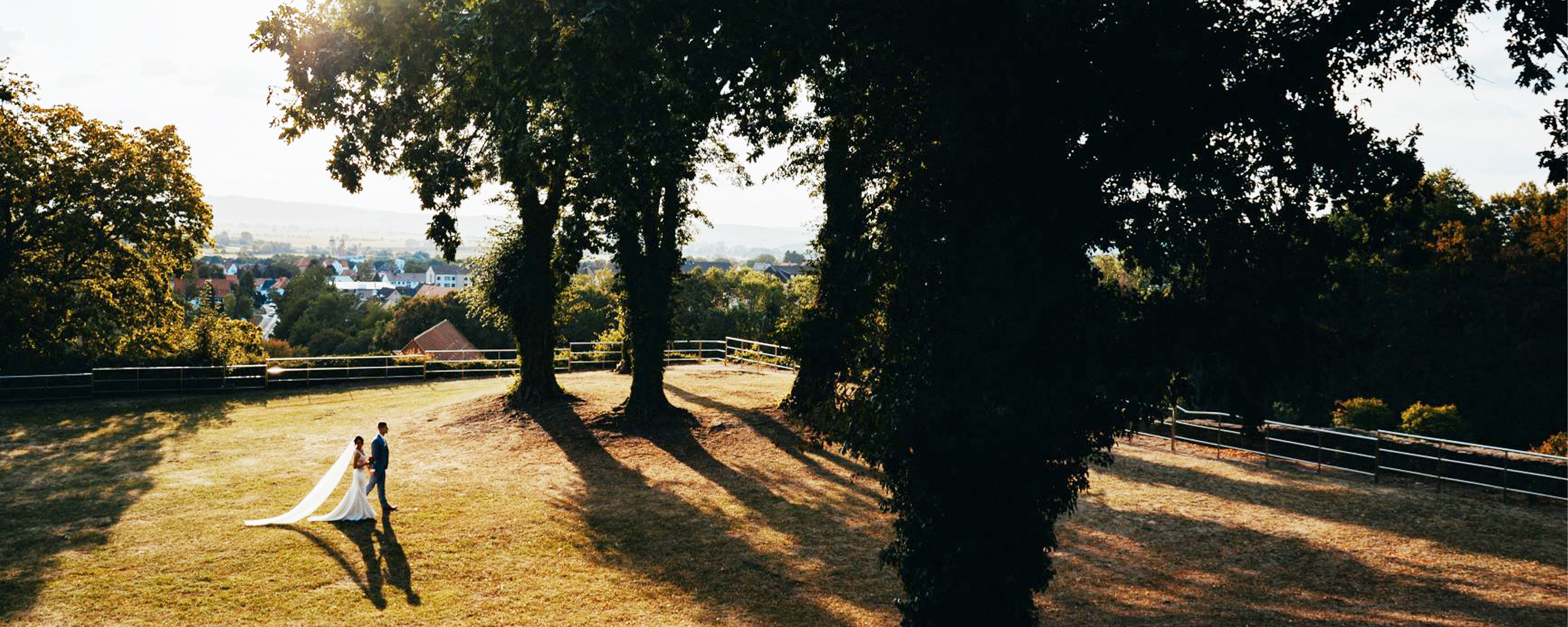 Brautpaar steht unter großen Bäumen auf einer sonnigen Wiese mit Blick auf eine Stadtlandschaft im Hintergrund. Die Braut trägt ein langes, weißes Kleid mit einem Schleier, der sich im Sonnenlicht abzeichnet, während der Bräutigam einen eleganten Anzug trägt.