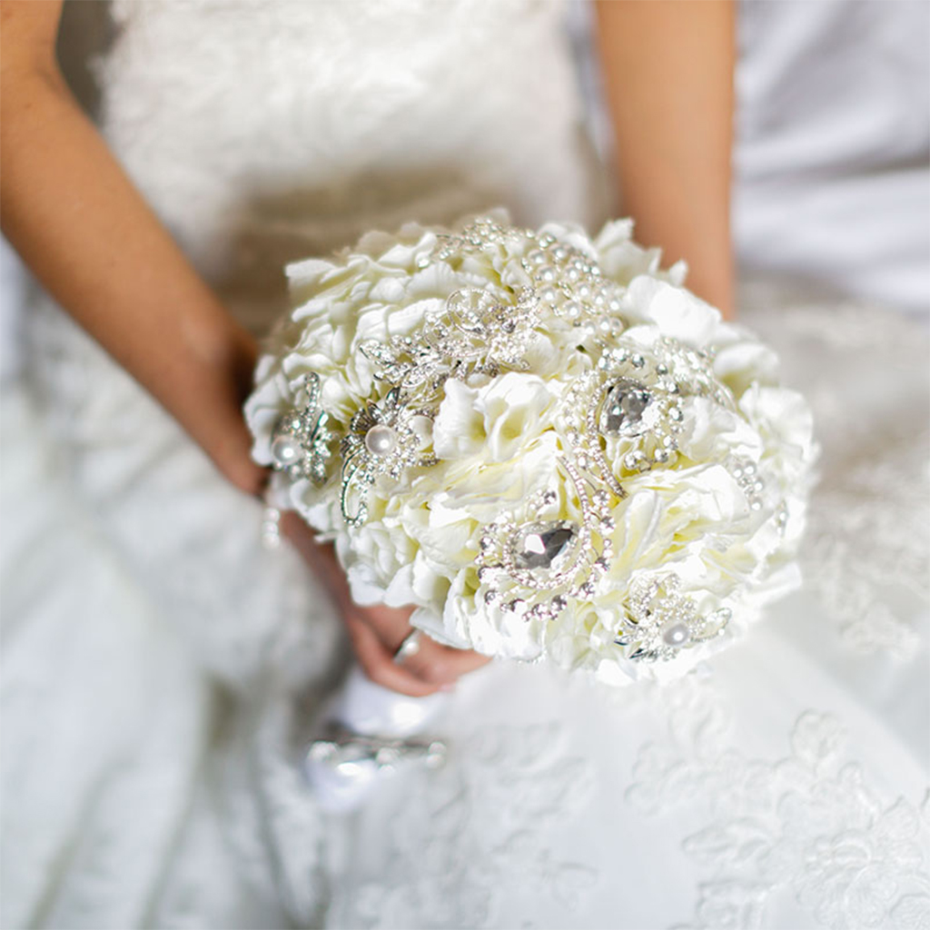 Eine Braut hält einen kunstvoll verzierten Hochzeitsstrauß aus weißen Blumen, geschmückt mit Perlen und funkelnden Strasssteinen. Dieses elegante Detail spiegelt die Schönheit und den Glanz einer klassischen Hochzeit wider, ideal für eine Hochzeit im Hotel.