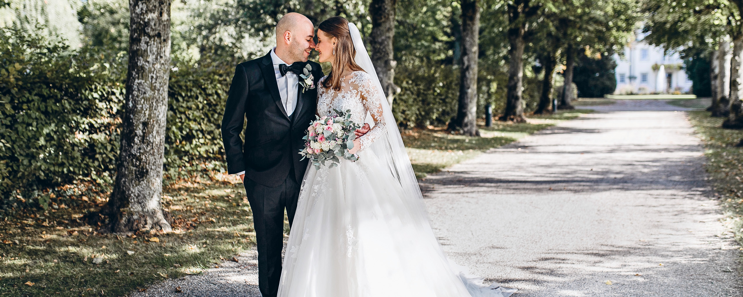 Ein Brautpaar steht eng umschlungen auf einem von Bäumen gesäumten Weg vor einer eleganten Villa. Die Braut trägt ein weißes Spitzenkleid und hält einen Blumenstrauß, während der Bräutigam in einem klassischen schwarzen Anzug gekleidet ist. Diese malerische Szene vermittelt Romantik und Eleganz, ideal für eine Hochzeit in einer exklusiven Location.