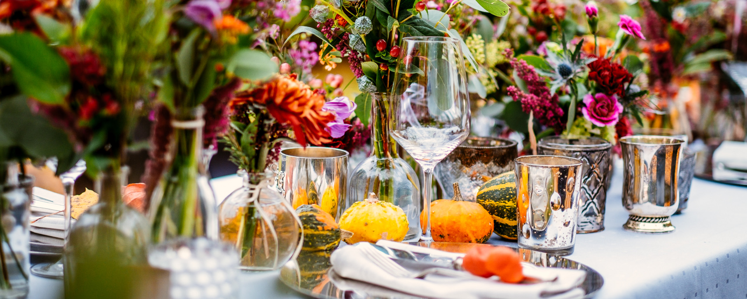 Farbenfrohe Tischdekoration für eine Hochzeit mit Blumenarrangements in verschiedenen Vasen, umgeben von eleganten Kerzenhaltern und herbstlichen Kürbissen. Die lebhafte und detailreiche Dekoration schafft eine festliche Atmosphäre für eine Hochzeit im Freien oder in einem stilvollen Veranstaltungsort.