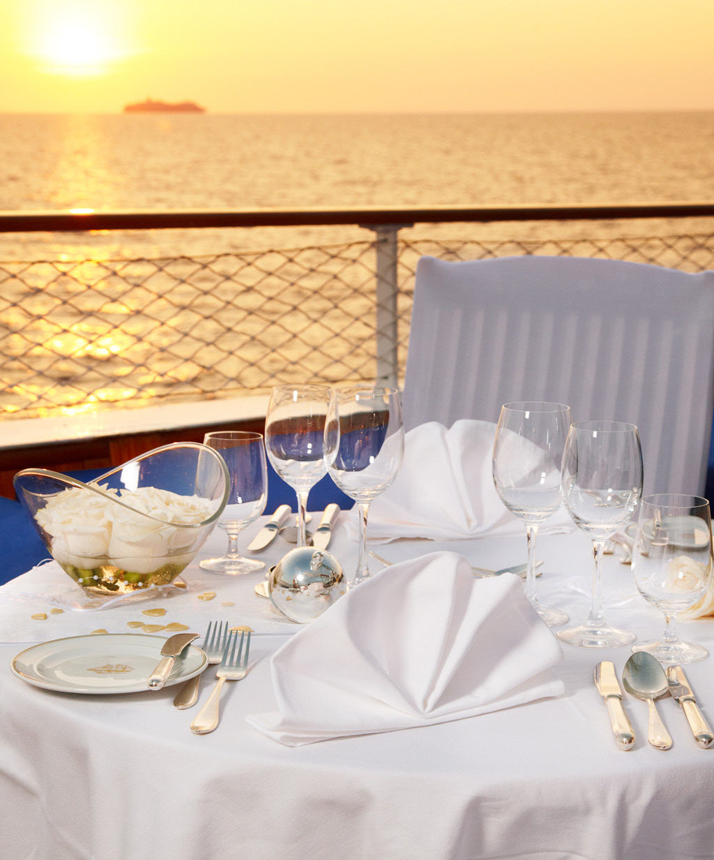 Romantisch gedeckter Tisch für eine Hochzeitsfeier auf einem Schiff, mit Blick auf den Sonnenuntergang über dem Meer. Die weiße Tischdekoration und die elegant arrangierten Gläser schaffen eine festliche Atmosphäre, die diese maritime Hochzeitslocation zu einem unvergesslichen Erlebnis macht.