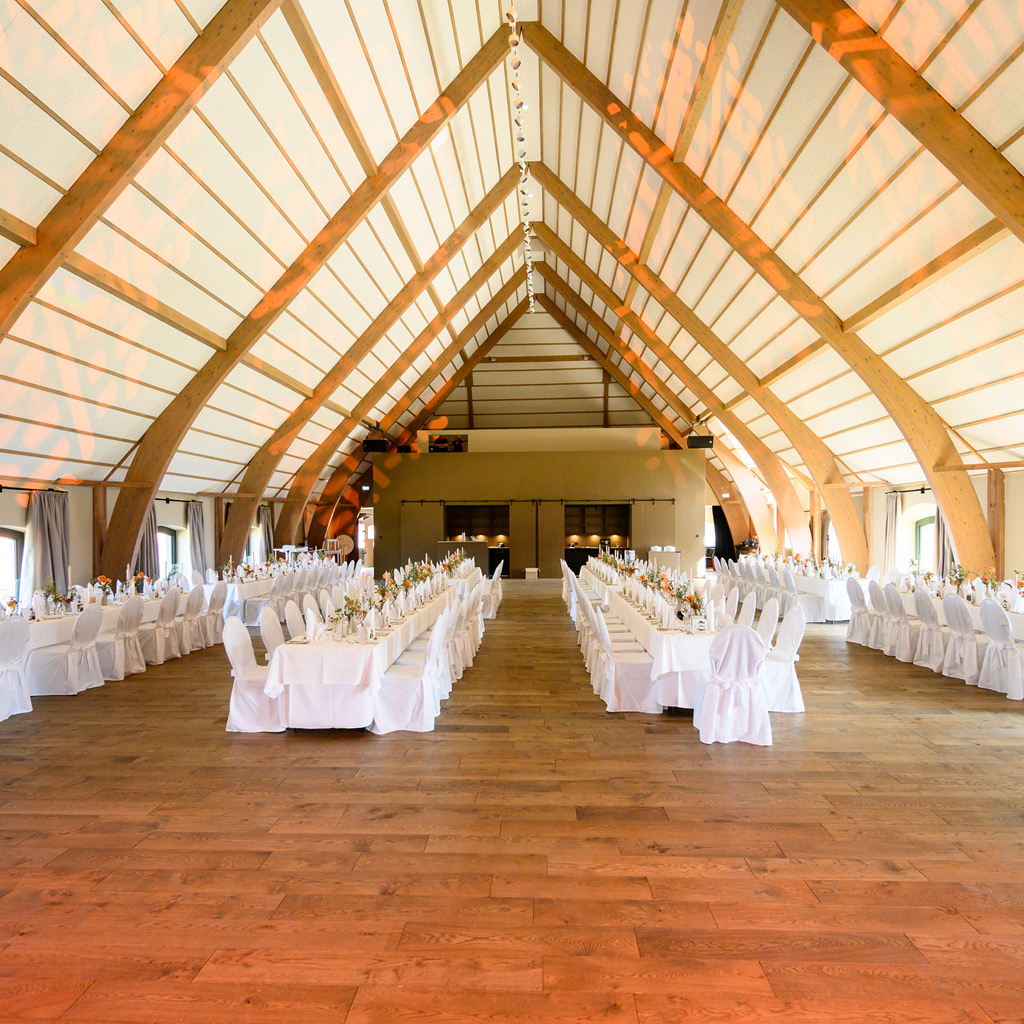 Großer Hochzeitssaal mit einer hohen, gewölbten Holzdecke, in dem lange, festlich gedeckte Tische mit weißen Tischdecken und Stühlen für eine Hochzeitsfeier vorbereitet sind. Die Tischdekoration besteht aus eleganten Blumenarrangements, und der Raum ist für den Empfang der Hochzeitsgäste bereit.