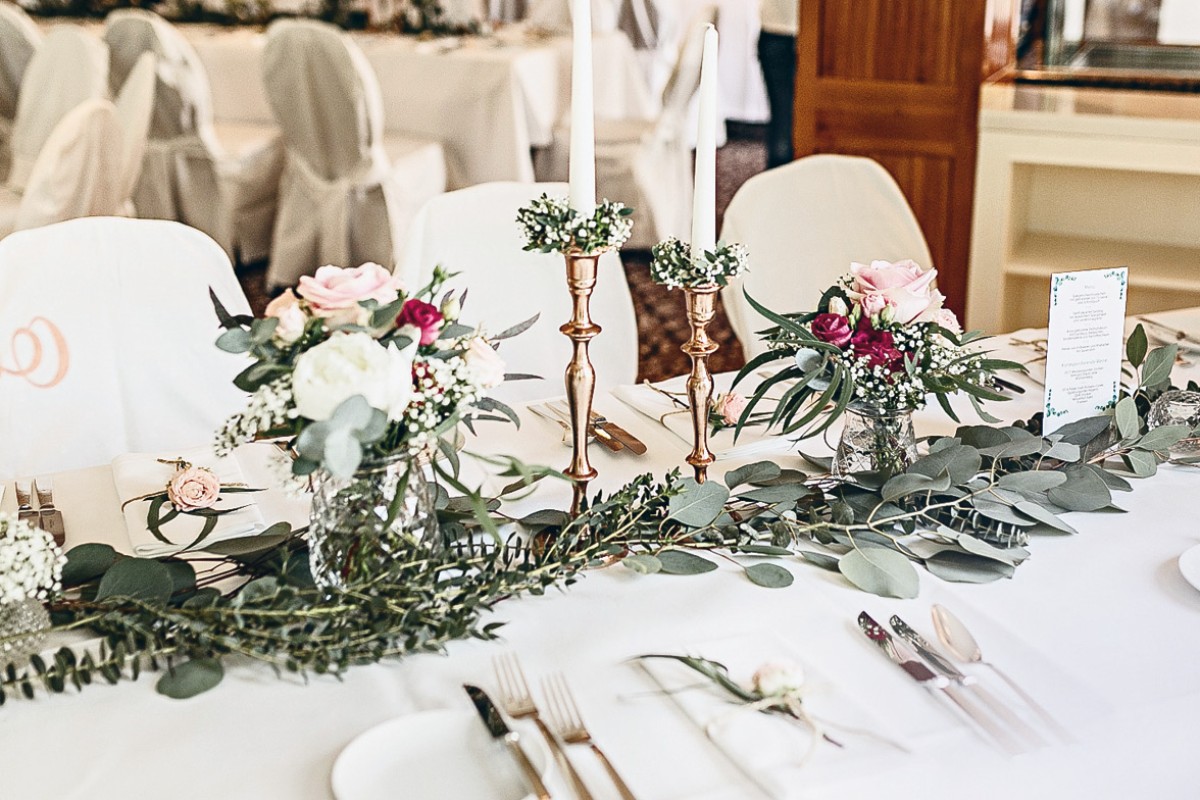 Festlich gedeckter Hochzeitstisch mit eleganter Blumendekoration aus Rosen und Eukalyptuszweigen. Auf dem Tisch befinden sich goldene Kerzenhalter mit weißen Kerzen, fein gedecktes Besteck und Menükarten, die für eine romantische und stilvolle Hochzeitsfeier vorbereitet sind.