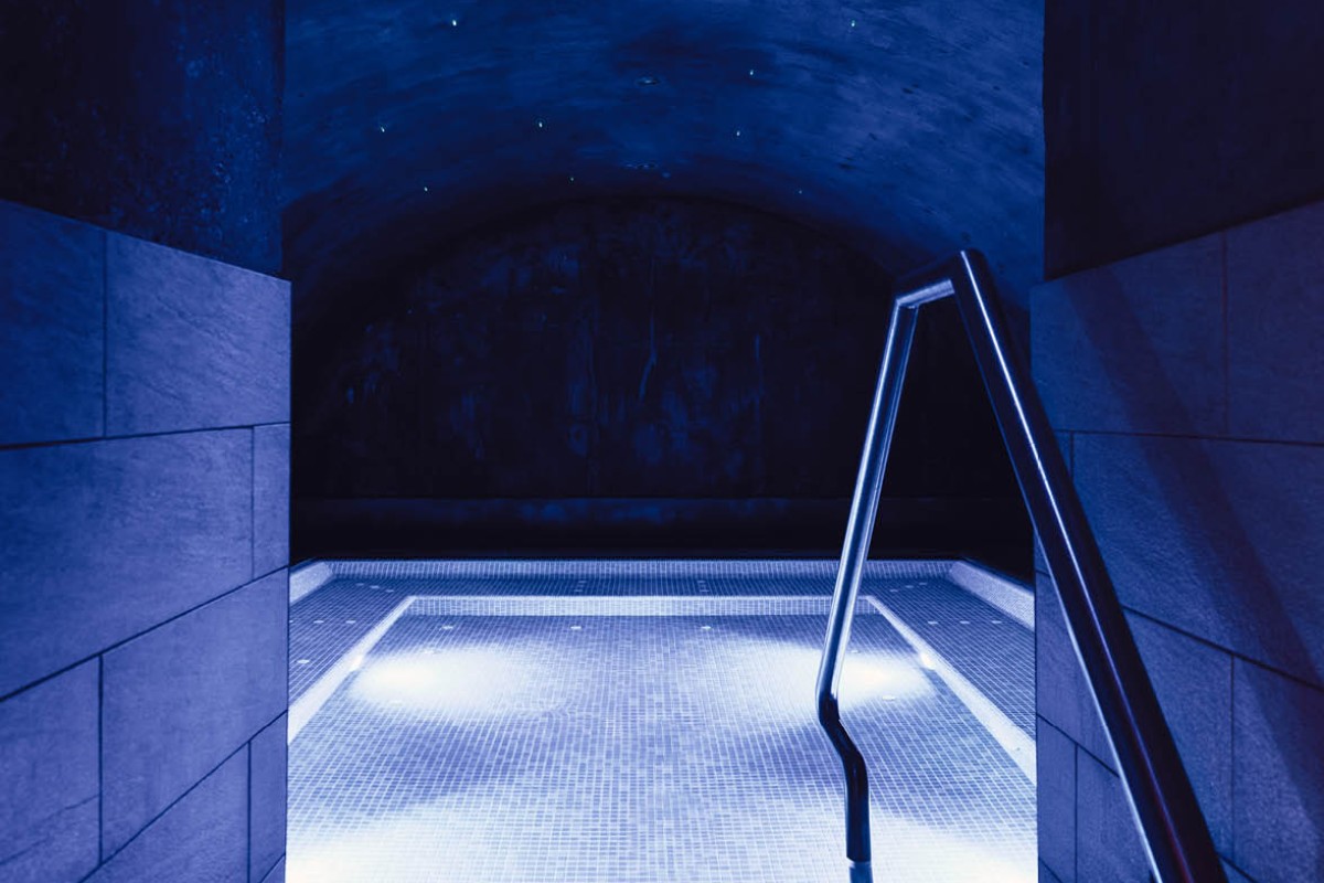 Das Bild zeigt ein stimmungsvolles, in blaues Licht getauchtes Spa-Becken in einer Höhle oder einem Raum mit gewölbter Decke. Eine Treppe führt ins Wasser, das in der Mitte sanft beleuchtet ist. Die Atmosphäre wirkt beruhigend und lädt zur Entspannung ein.