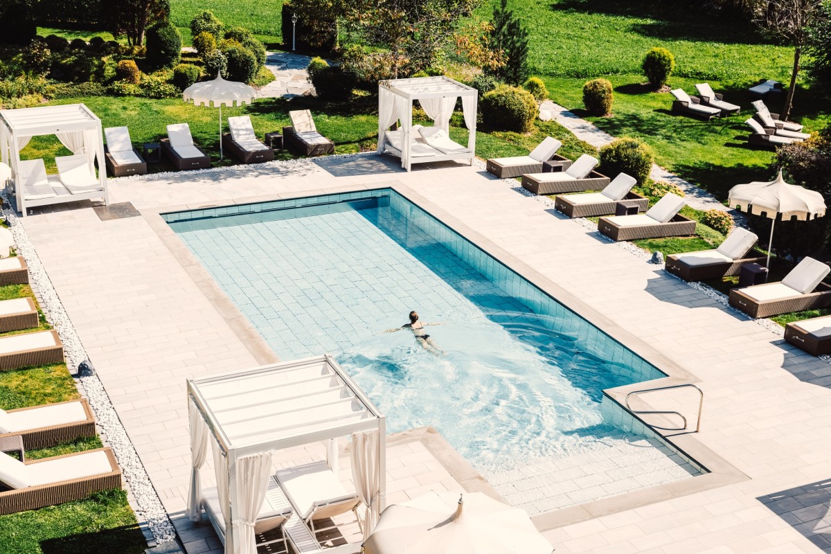 Das Bild zeigt einen eleganten Außenpool in einem gepflegten Garten eines Wellnesshotels, umgeben von luxuriösen Sonnenliegen und gemütlichen Cabanas. Eine Person genießt eine entspannende Schwimmrunde im klaren Wasser, während die sonnige Umgebung für eine perfekte Erholung sorgt.