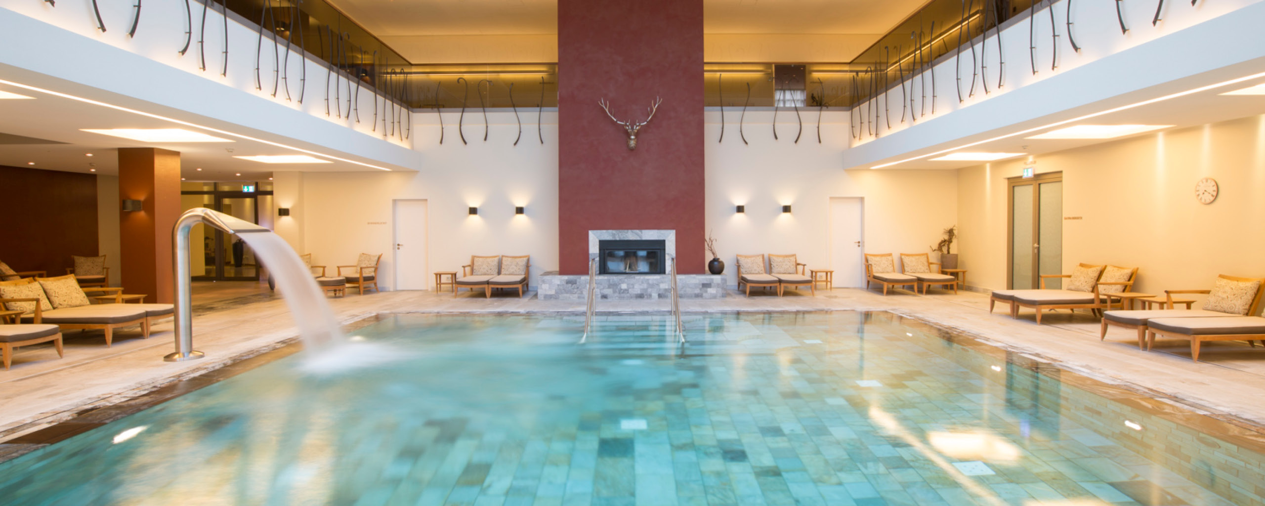 Ein luxuriöser Innenpool in einem Wellnesshotel, umgeben von eleganten Liegestühlen und einer entspannenden Atmosphäre. Das moderne Design, kombiniert mit der warmen Beleuchtung und einem Kamin, bietet Gästen eine ruhige Oase für Erholung und Entspannung.