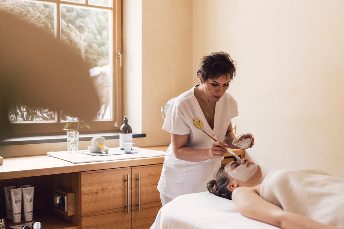 Das Bild zeigt eine entspannte Wellnessbehandlung in einem Spa. Eine professionelle Kosmetikerin trägt eine Gesichtsmaske bei einer Kundin auf, die ruhig auf einer bequemen Liege liegt. Der Raum ist in warmen, natürlichen Tönen gehalten, was eine beruhigende und friedliche Atmosphäre schafft. Im Hintergrund ist ein Fenster zu sehen, das den Raum mit natürlichem Licht erhellt und einen Blick auf die Außenwelt bietet. Produkte und Utensilien, die zur Pflege und Entspannung beitragen, sind auf einer Holzkommode arrangiert, was den Raum noch einladender macht.