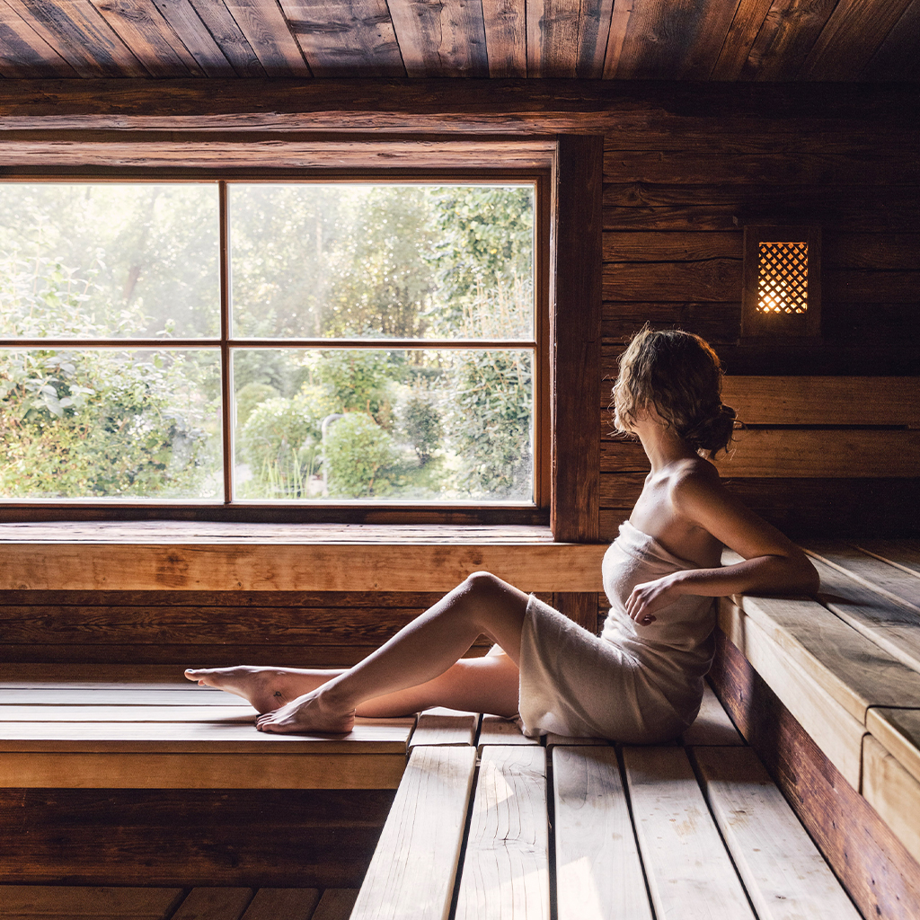 Eine Frau entspannt in einer holzverkleideten Sauna und genießt den Blick auf die grüne, friedliche Natur durch ein großes Fenster. Diese Szene verkörpert die Ruhe und Erholung, die ein luxuriöses Spa in einem erstklassigen Wellnesshotel bietet.